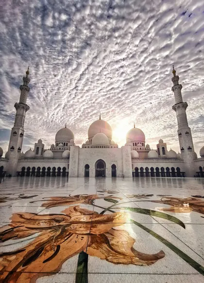 Una moschea bianca con cupole e minareti riflessi su un cortile lucido con un grande mosaico floreale, sotto un cielo nuvoloso.