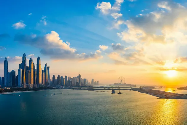 Una vista aerea dello skyline di una città moderna con grattacieli in riva al mare, illuminata da un tramonto dorato.