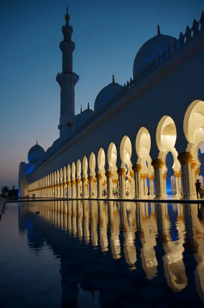 Una moschea bianca illuminata con un colonnato di archi e un minareto che si riflette nell'acqua calma al crepuscolo.