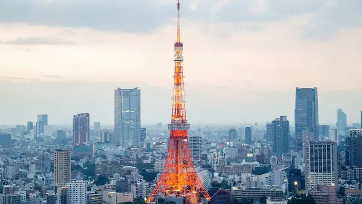 Una torre reticolare illuminata rossa e bianca si erge imponente tra un vasto skyline cittadino ricco di edifici moderni sotto un cielo nuvoloso.
