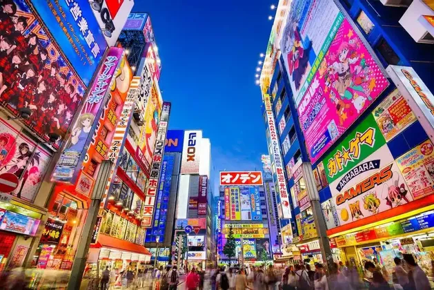 Una trafficata strada cittadina al tramonto è affiancata da alti edifici coperti da luminosi e colorati cartelloni e pubblicità giapponesi.