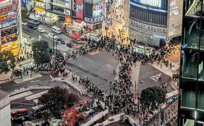 Una vista aerea di un vivace incrocio cittadino di notte, dove folle di persone con effetto mosso attraversano le strade tra alti edifici illuminati.