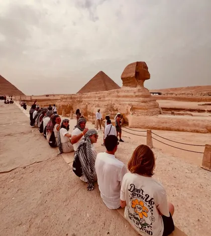 Un gruppo WeRoad in fila indiana su una sporgenza rocciosa, ammira la Grande Sfinge e le piramidi sotto un cielo coperto.