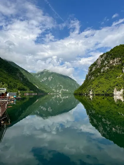 Montagne ripide e verdi e un cielo parzialmente nuvoloso si riflettono nelle acque calme di un lago.