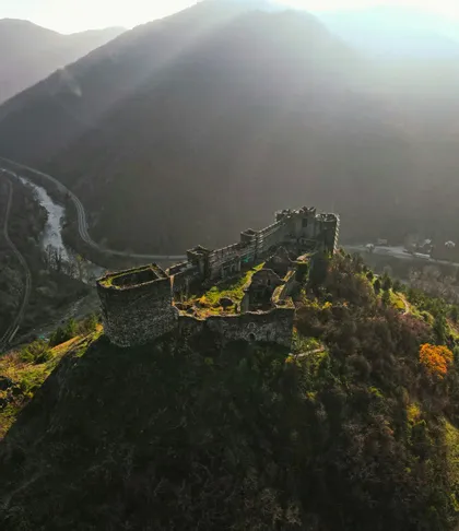 Una vista aerea delle rovine di un castello in pietra su una ripida collina boscosa che si affaccia su una valle fluviale, con il sole che spunta sulle montagne lontane.