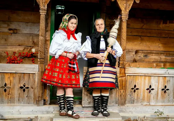 Due donne in abiti tradizionali su un portico di legno, una con una conocchia per filare la lana.