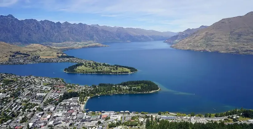 Vista aerea di una cittadina lacustre e isolotti su un grande lago blu, con una catena montuosa scoscesa sullo sfondo.
