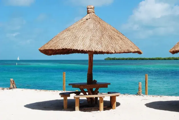 Un tavolo da picnic in legno con panche si trova sotto un grande ombrellone di paglia, su una spiaggia di sabbia bianca con vista su un oceano turchese.