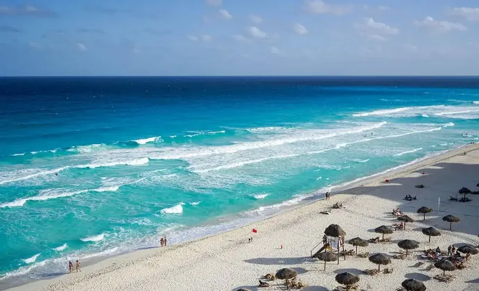 Vista aerea di una spiaggia di sabbia bianca con acqua turchese dove le persone si rilassano sotto ombrelloni di paglia.