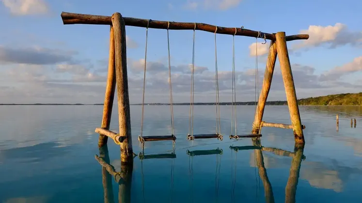 Un'altalena di legno, con diversi seggiolini vuoti, si erge su un'acqua calma e limpida, il suo riflesso visibile in superficie.