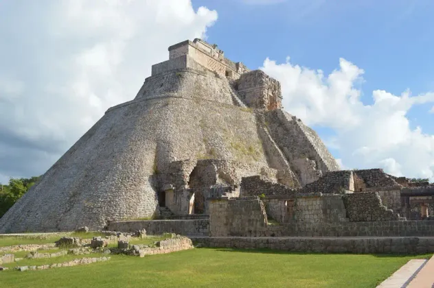 Una grande antica piramide di pietra con angoli arrotondati si erge su un campo erboso con altre rovine sotto un cielo parzialmente nuvoloso.
