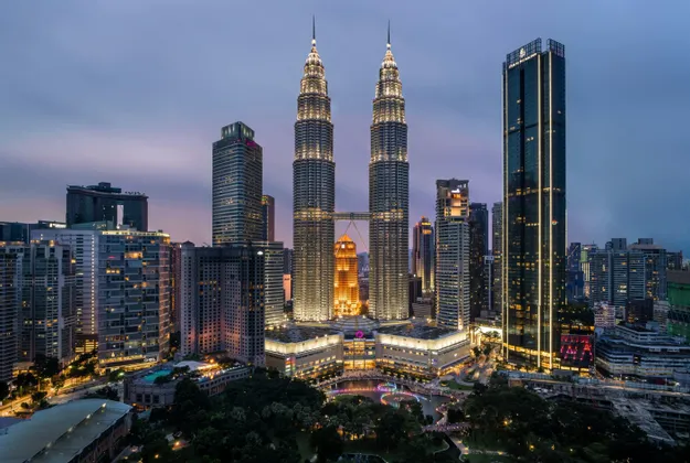 Due alti grattacieli gemelli illuminati spiccano nello skyline di una città moderna al tramonto, circondati da altri edifici illuminati.