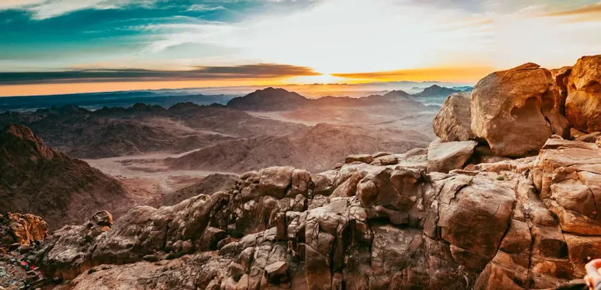 The sun rises over a vast, hazy mountain range, viewed from a rocky summit under a turquoise and orange sky.