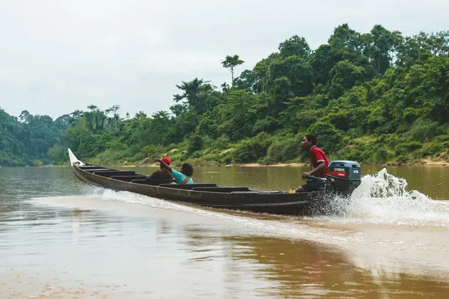 Tre persone su un lungo motoscafo di legno navigano lungo un fiume, con una che indica la fitta giungla sulla riva.