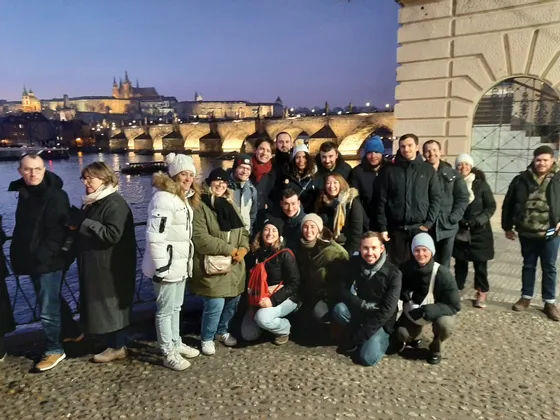Un groupe WeRoad prend la pose pour une photo sur une promenade pavée au crépuscule, avec un pont illuminé et un panorama urbain de l'autre côté de la rivière en arrière-plan.