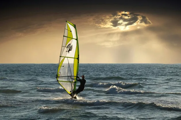 Un windsurfer solca le onde agitate dell'oceano con una vela verde e bianca, sotto un tramonto nuvoloso e spettacolare.