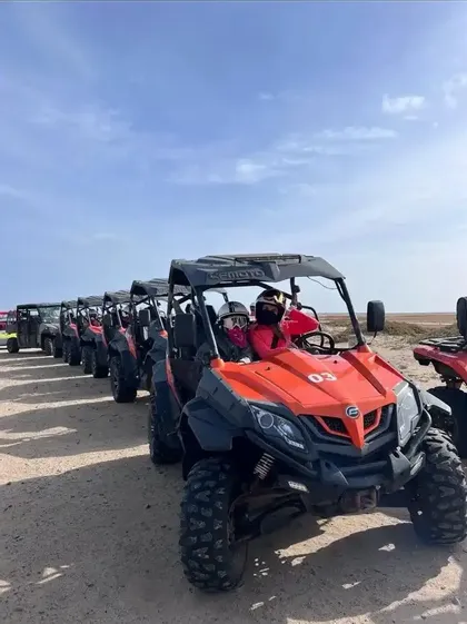 Un viaggio di gruppo WeRoad con una lunga fila di buggy fuoristrada arancioni su un sentiero sabbioso sotto un cielo azzurro limpido.