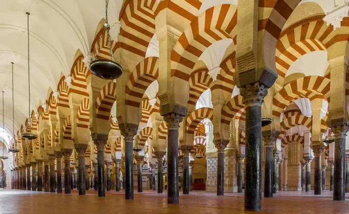 Fila di colonne scure sorreggono doppi archi a strisce rosse e bianche in una grande sala con un alto soffitto a volta e lampade a sospensione.