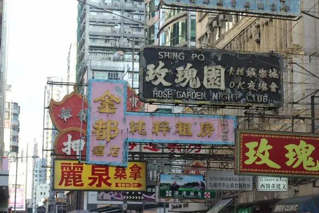 Un fitto gruppo di insegne logore con caratteri cinesi pende su una strada urbana davanti a edifici alti.