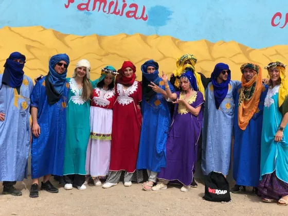 Un grupo de WeRoad en viaje, ataviado con coloridas túnicas tradicionales y pañuelos en la cabeza, posa para una foto frente a una pared del desierto pintada.