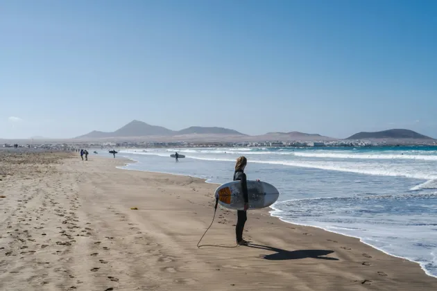 Un surfista in muta tiene una tavola da surf su una spiaggia sabbiosa, guardando l'oceano con montagne distanti sotto un cielo sereno.