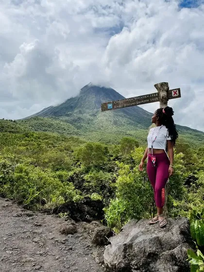 Eine Frau auf einem Wanderweg steht auf einem Felsen und blickt auf einen großen, teilweise von Wolken bedeckten Vulkan.
