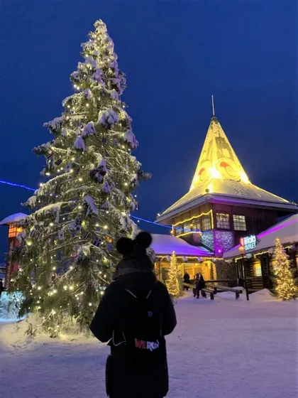 Eine Person mit WeRoad Rucksack betrachtet nachts auf einem Dorfplatz einen großen, schneebedeckten und beleuchteten Weihnachtsbaum.