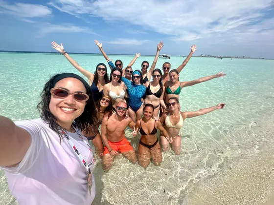 Un groupe WeRoad souriant pour un selfie, les pieds dans l'eau turquoise peu profonde et claire.