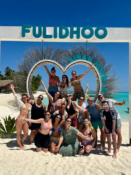 Eine WeRoad Gruppenreise posiert gemeinsam für ein Foto an einem weißen Sandstrand vor einem herzförmigen Rahmen unter strahlend blauem Himmel.