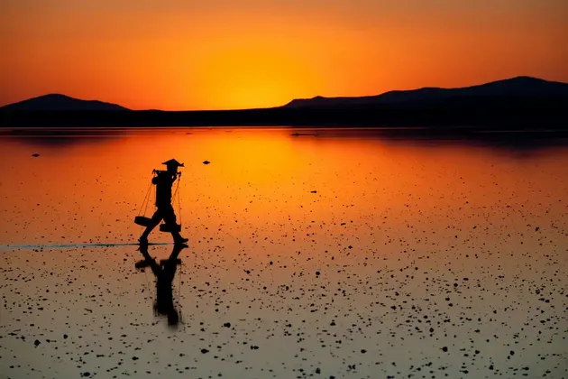 Una sagoma con cappello conico e bilanciere in spalla cammina in acque basse e riflettenti al tramonto.