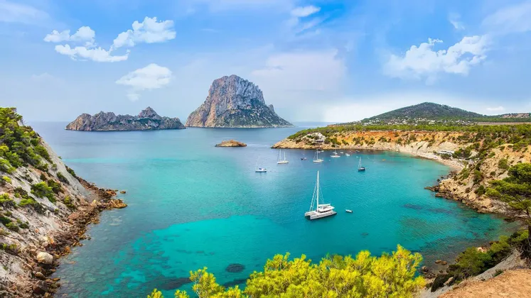 Barche a vela e un catamarano sono ancorati in una baia calma e turchese, circondata da scogliere rocciose e una grande isola.