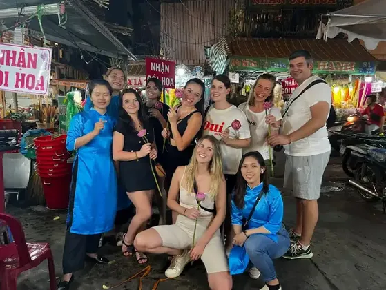 Un viaggio di gruppo WeRoad posa per una foto in un vivace mercato notturno, con diversi membri che tengono in mano fiori di loto rosa.