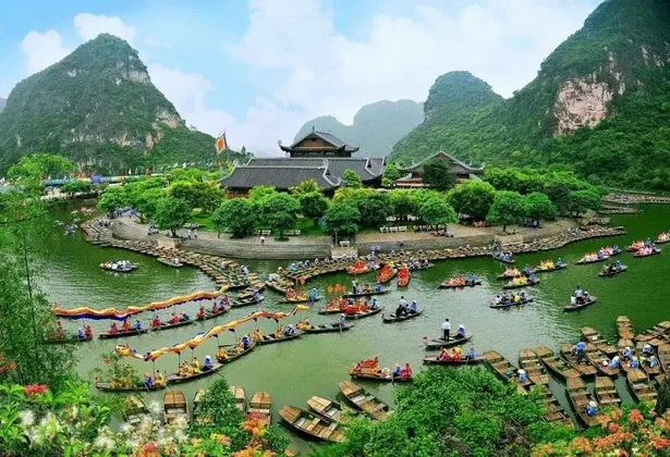 Decine di imbarcazioni tradizionali piene di gente navigano su un fiume che circonda un complesso di templi ai piedi di lussureggianti montagne verdi.