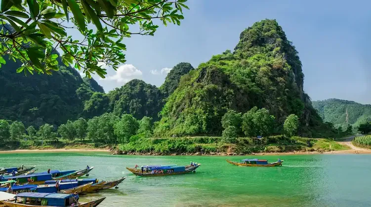 Barche lunghe tradizionali su un fiume di un verde vivace, ai piedi di lussureggianti montagne ricoperte di foreste, sotto un cielo soleggiato.