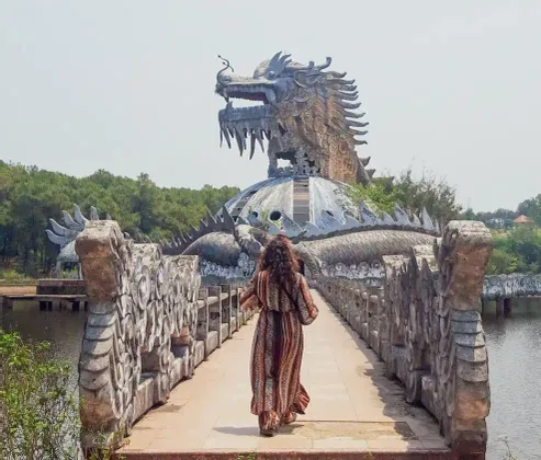 Una donna, vista di spalle, cammina su un ponte di pietra in direzione di una grande scultura di drago, logorata dal tempo e costruita su un lago.