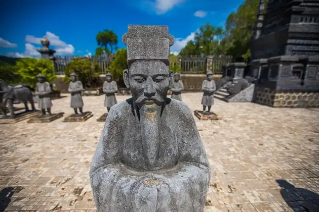 Una statua in pietra consumata di un uomo in abiti tradizionali si erge in un cortile pavimentato con altre statue sullo sfondo.