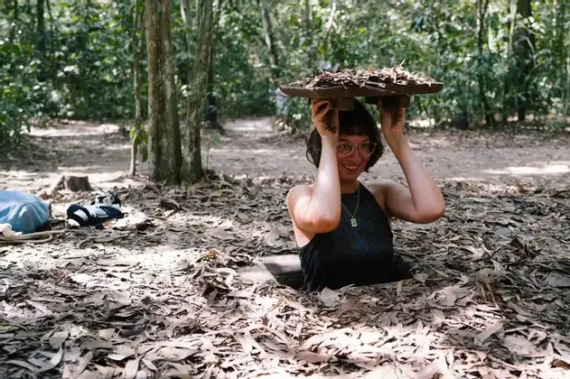 Una donna sorridente con gli occhiali emerge da una buca ricoperta di foglie in una foresta, reggendo il coperchio di foglie sopra la testa.