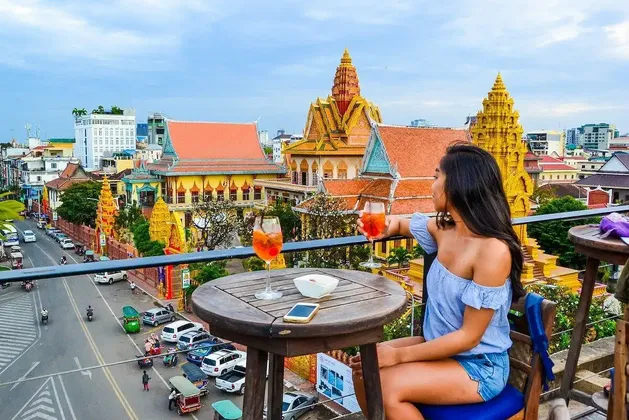 Una donna siede a un tavolo su un balcone sul tetto con due cocktail, affacciata su una città con templi dorati e traffico stradale.