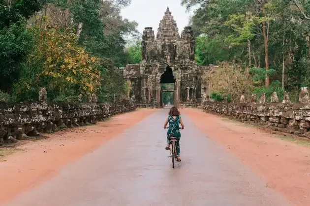 Una persona vista di spalle pedala in bicicletta su una strada che porta a un grande, antico portale in pietra affiancato da alberi della giungla.