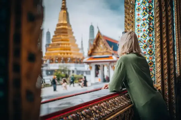 Una donna bionda con una camicia verde si sporge da un balcone decorato, ammirando un tempio dorato.