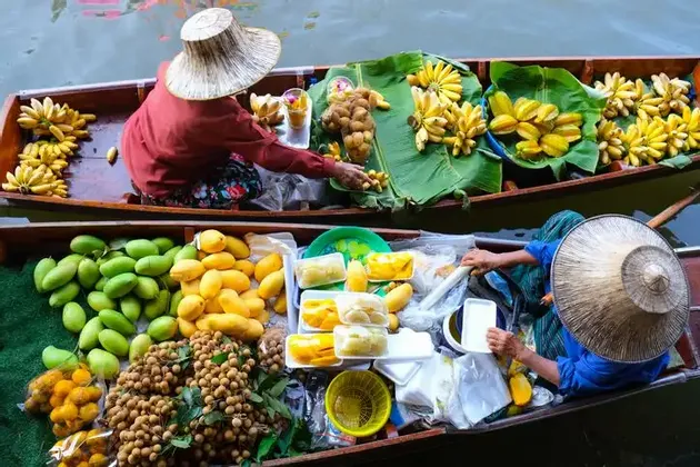 Vista aerea di due venditori con cappelli a cono che offrono frutta fresca, come banane e manghi, dalle loro lunghe barche di legno in un mercato galleggiante.
