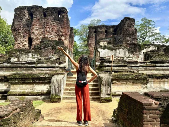 Une femme, sac à dos, vue de dos, le bras levé devant d'anciennes ruines de briques moussues.