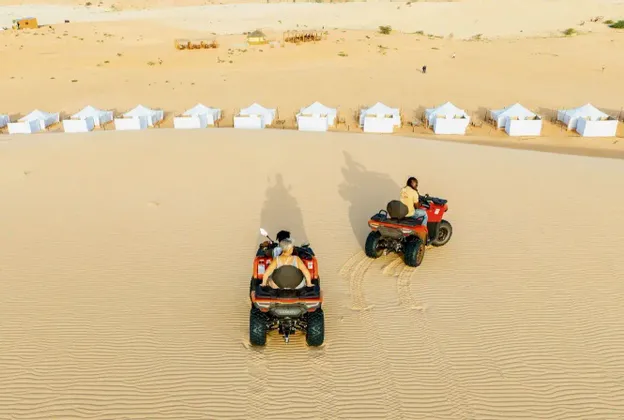 Vista aérea de dos personas de un viaje en grupo de WeRoad en quads por dunas de arena, con un campamento en el desierto visible al fondo.
