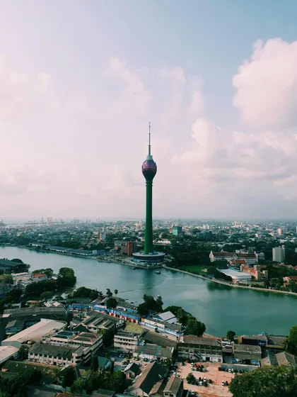Una torre verde e alta con la cima a forma di loto si erge al centro di uno scenario urbano, accanto a un grande specchio d'acqua.