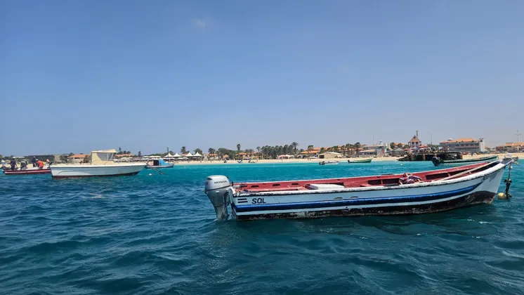Una lancha motora roja y blanca flota en aguas azules, con una playa de arena y un pueblo costero de fondo.