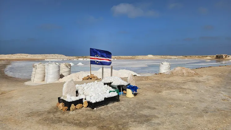 La bandera de Cabo Verde se alza entre pilas de sal cosechada en sacos y cajas en una salina bajo un cielo azul.