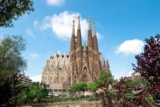 An ornate basilica with multiple tall, intricate spires stands behind a park with a pond and trees under a partly cloudy blue sky.