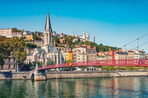A red footbridge crosses a river, leading to a historic European city with a church and basilica on a hill against a clear sky.