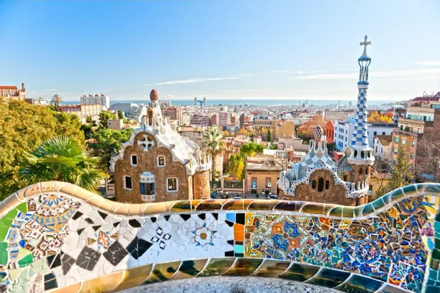 A colorful mosaic bench overlooks whimsical buildings and a cityscape stretching to the sea under a clear blue sky.