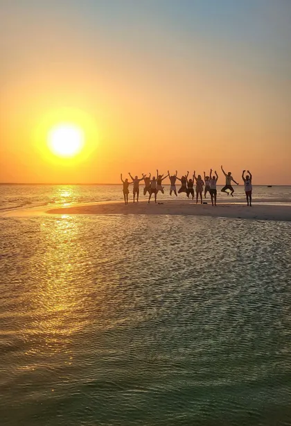 Un viaje en grupo de WeRoad salta sobre un banco de arena, silueteado contra el atardecer sobre el agua.